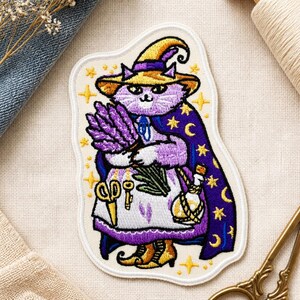 May include: An embroidered patch featuring a purple cat wearing a yellow pointed hat and a dark blue cape with yellow stars and moons. The cat holds a bundle of lavender and wears a white apron with a pair of gold scissors, a key, and a potion bottle.