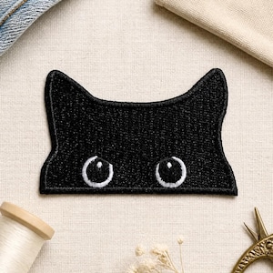 Puede incluir: Un parche bordado negro con forma de parte superior de cabeza de gato con dos orejas puntiagudas. El parche presenta dos ojos circulares bordados en blanco con pupilas negras, creando un efecto de asomo. Los bordes tienen costuras negras.