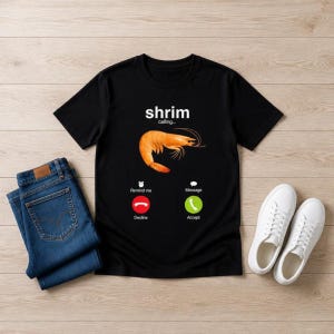 May include: A black t-shirt featuring a graphic of an orange shrimp and the text 'shrim calling...' above it. Below the shrimp are icons for 'Remind me', 'Message', a red decline call button, and a green accept call button.