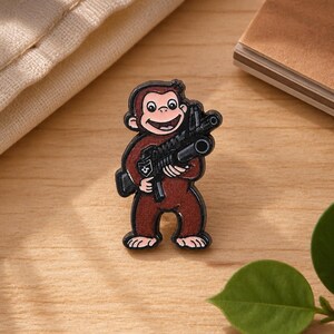 May include: A brown enamel pin featuring a cartoon monkey with a light pink face, hands, and feet, holding a black rifle. The monkey has a smiling expression with visible teeth and is outlined in black metal, creating a distinct character design.