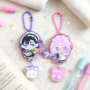 Könnte beinhalten: Zwei Schlüsselanhänger im Anime-Stil mit Dienstmädchen-Charakteren. Ein Schlüsselanhänger ist lila mit einem schwarzhaarigen Dienstmädchen, das Essen hält, und der andere ist rosa mit einem pinkhaarigen Dienstmädchen, das einen Besen hält. Jeder Schlüsselanhänger hat einen passenden Anhänger und eine Kugelkette.