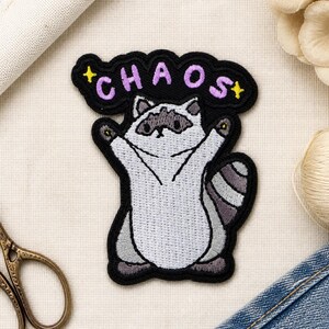 Op de afbeelding: Een geborduurde patch met een lichtgrijze wasbeer met donkergrijze oogvlekken en een gestreepte staart, staand met de armen omhoog. Boven de wasbeer staat het woord CHAOS in paarse letters op een zwarte achtergrond met twee gele sterren.