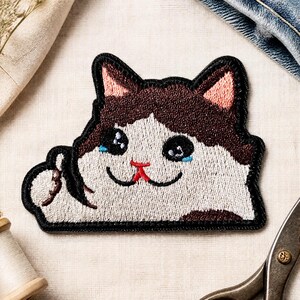 Könnte beinhalten: Ein gestickter Aufnäher, der eine braun-weiße Katze mit großen, glänzenden Augen und kleinen blauen Tränen zeigt. Die Katze hat rosafarbene Innenohren, eine rote Nase und eine schwarze Umrandung. Eine Pfote ist in Gesichtsnähe erhoben.