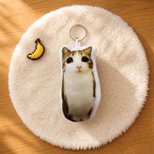 May include: A plush keyring featuring a brown and white tabby cat with large dark eyes, accompanied by a small pixelated yellow banana charm. The cat keyring has a silver metal ring attached to the top for hanging or attachment purposes.