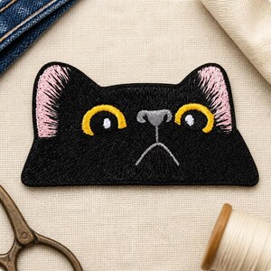 Op de afbeelding: Een geborduurde patch in de vorm van een zwarte kattenkop met grote gele ogen, witte pupillen, een grijze neus en roze binnenoren. De kat heeft een rechte, neutrale mondlijn en is uitgevoerd in zwart garen op een textiele ondergrond.