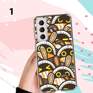 Puede incluir: Una funda transparente para tel&eacute;fono con un patr&oacute;n repetido de caras de conejos de dibujos animados en marr&oacute;n, naranja y blanco. La funda tiene un borde transparente y un recorte para la c&aacute;mara. La funda del tel&eacute;fono la sostiene una persona.