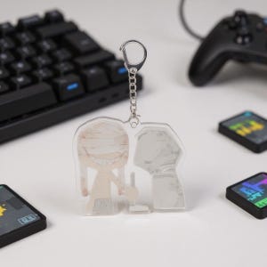 Puede incluir: Un llavero acr&iacute;lico transparente con un personaje de dibujos animados con vendas y una amplia sonrisa, junto con una figura sombreada. El llavero cuelga de una cadena y un cierre plateados. Un teclado negro, un mando de juegos y cartuchos de juegos est&aacute;n en el fondo.