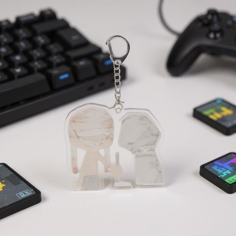 Puede incluir: Un llavero acr&iacute;lico transparente con un personaje de dibujos animados con vendas y una amplia sonrisa, junto con una figura sombreada. El llavero cuelga de una cadena y un cierre plateados. Un teclado negro, un mando de juegos y cartuchos de juegos est&aacute;n en el fondo.