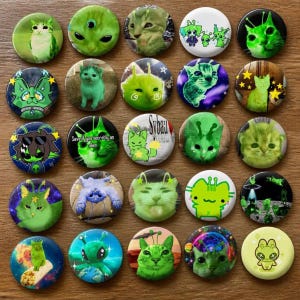 Op de afbeelding: Een verzameling van vijfentwintig ronde buttons met diverse illustraties van groene katten met buitenaardse kenmerken, inclusief antennes en ruimtethema-achtergronden. Sommige buttons bevatten teksten zoals Sybau en Save if you secretly an alien.