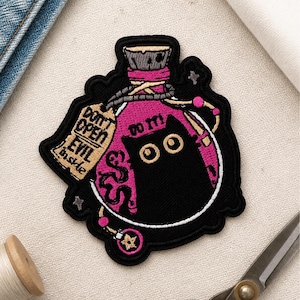 Puede incluir: Un parche bordado que muestra una botella de poción rosa con la silueta de un gato negro en su interior. La botella tiene un tapón de corcho, una etiqueta beige con el texto "DON'T OPEN EVIL INSIDE" y la frase "DO IT!" sobre el gato.