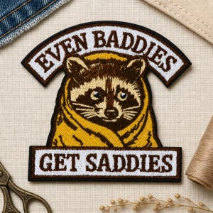 May include: An embroidered patch featuring a brown and white raccoon wrapped in a yellow blanket. The patch has a curved top banner with the text EVEN BADDIES and a rectangular bottom banner with the text GET SADDIES in brown lettering.
