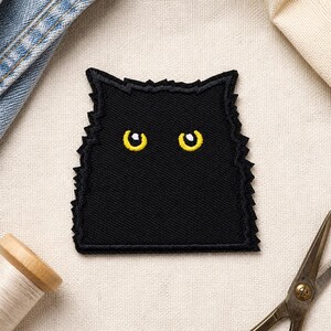 May include: A black embroidered patch shaped like a cat head with jagged edges and two bright yellow eyes. The patch features a dense, textured thread construction with a distinct outline, showcasing a stylised feline face in a dark, solid colour.