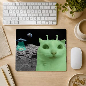 以下が含まれることがあります： 頭に2本のアンテナをつけたライトグリーンの猫が月面風景の中に描かれた正方形のマウスパッド。空飛ぶ円盤が月面に緑色の光線を放っており、背景の暗い宇宙空間には地球が見えている。