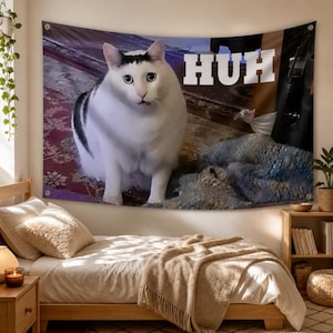 Könnte beinhalten: Ein rechteckiger Wandteppich mit einem großen weißen Katzenmotiv, das schwarze Abzeichen am Kopf und Rücken aufweist und auf einem gemusterten Boden sitzt. Das Wort HUH ist in großen weißen Blockbuchstaben oben rechts auf dem Stoff gedruckt.