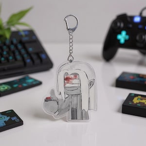 Puede incluir: Un llavero acr&iacute;lico transparente con un personaje de dibujos animados con cabello blanco, una venda sobre los ojos y un bot&oacute;n rojo. El llavero est&aacute; sujeto a una cadena y un cierre plateados. El fondo incluye un teclado, un mando de juegos y cartuchos de juegos.