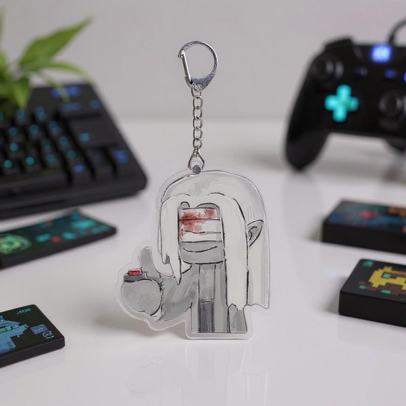 Puede incluir: Un llavero acr&iacute;lico transparente con un personaje de dibujos animados con cabello blanco, una venda sobre los ojos y un bot&oacute;n rojo. El llavero est&aacute; sujeto a una cadena y un cierre plateados. El fondo incluye un teclado, un mando de juegos y cartuchos de juegos.