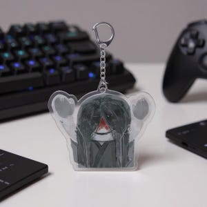 Puede incluir: Un llavero acr&iacute;lico transparente con una ilustraci&oacute;n estilizada de una figura con pelo largo negro y una marca roja. El llavero tiene una cadena y un cierre plateados. El dise&ntilde;o est&aacute; sobre un fondo borroso con un teclado y un mando de juegos.