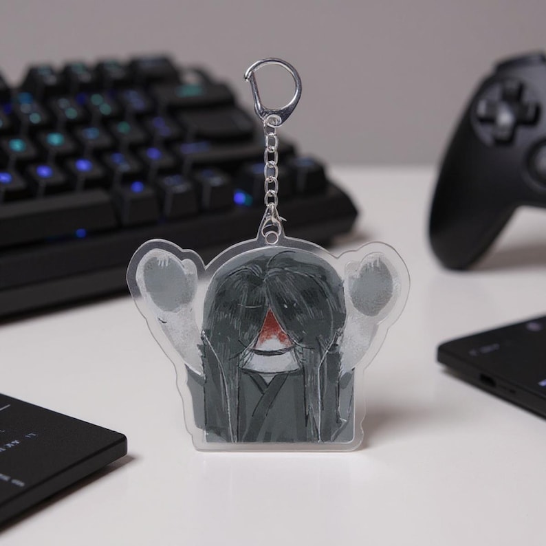 Puede incluir: Un llavero acr&iacute;lico transparente con una ilustraci&oacute;n estilizada de una figura con pelo largo negro y una marca roja. El llavero tiene una cadena y un cierre plateados. El dise&ntilde;o est&aacute; sobre un fondo borroso con un teclado y un mando de juegos.