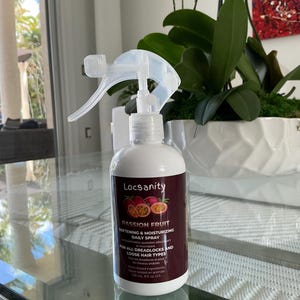 Op de afbeelding: Een doorzichtige plastic fles met een spuitmond, met het label "Locsanity Passion Fruit Softening & Moisturizing Daily Spray". De fles staat op een glazen tafel met een witte orchidee op de achtergrond. Het label vermeldt ook "Voor alle dreadlocks en los haar".