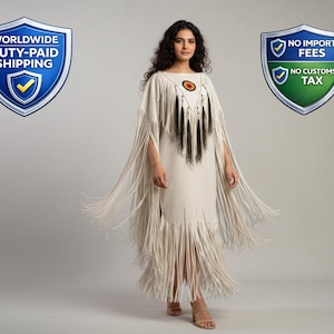May include: A cream-coloured dress with long fringe detailing. The dress features a round neckline with a circular design and black fringe accents. The model is wearing sandals. The dress is a unique, bohemian style.