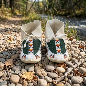 May include: A pair of cream-coloured leather moccasins featuring a dark green beaded vamp with a geometric pattern in white, orange, and yellow. The footwear includes matching cream leather laces tied at the front and a soft, flexible construction.