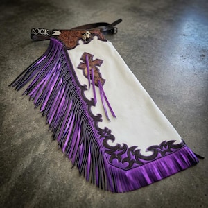 May include: A white leather chaps piece featuring a brown leather waistband with the word ROCK embossed, floral tooling, and a skull accent. The leg section is adorned with a brown leather cross overlay and long, metallic purple fringe trim.