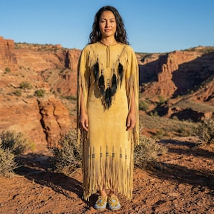 May include: A long, mustard-yellow dress with fringe detailing and black feather accents. The dress features a high neckline and long sleeves with fringe. The model is wearing matching mustard-yellow moccasins with blue and yellow beadwork. The background is a desert landscape.