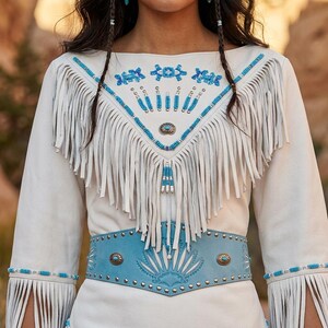 May include: Cream-colored suede dress with turquoise and silver embellishments. The dress features fringe detailing on the bodice and sleeves, a turquoise belt with metal studs, and intricate beadwork around the neckline and cuffs. The dress has a bohemian, Southwestern style.