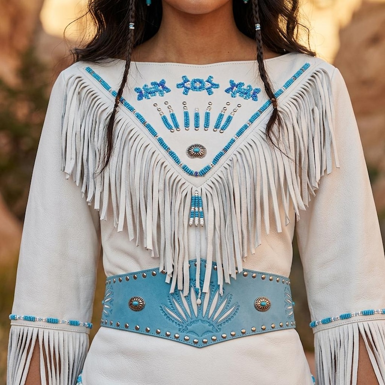 May include: Cream-colored suede dress with turquoise and silver embellishments. The dress features fringe detailing on the bodice and sleeves, a turquoise belt with metal studs, and intricate beadwork around the neckline and cuffs. The dress has a bohemian, Southwestern style.