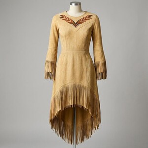 May include: A tan suede dress featuring long sleeves with fringe trim at the cuffs, a high-low hemline with extensive fringe detailing, and a V-shaped neckline adorned with a geometric pattern in brown, black, and white beads and thread.