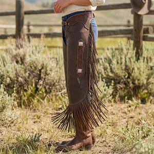 May include: Brown leather chaps with decorative tooling and fringe. The chaps feature silver-toned accents and are worn over blue jeans. The chaps are paired with brown cowboy boots, creating a Western-inspired look.