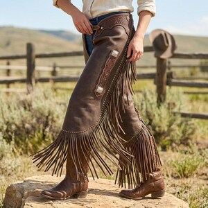 May include: Brown leather chaps with long fringe and decorative silver-toned studs and accents. The chaps are worn with brown cowboy boots. The image is set outdoors with a wooden fence and a natural background.