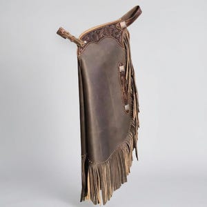 May include: Brown leather chaps with decorative tooling and fringe. The chaps feature a belt and buckle closure, silver-toned accents, and a detailed floral design. The fringe is a lighter brown color.