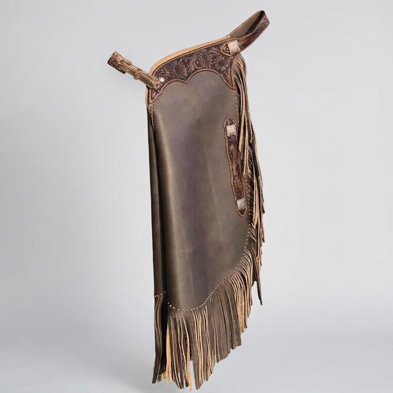 May include: Brown leather chaps with decorative tooling and fringe. The chaps feature a belt and buckle closure, silver-toned accents, and a detailed floral design. The fringe is a lighter brown color.