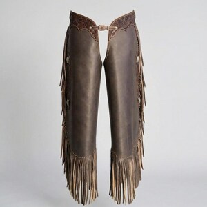 May include: Brown leather chaps with decorative tooling along the top and fringe along the sides and bottom. The chaps have a buckle closure at the waist and are designed to be worn over pants. The chaps are a western wear item.