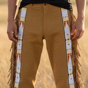 Puede incluir: Pantalones de ante color canela con rayas blancas con cuentas en los laterales. Las rayas presentan patrones geométricos rojos, amarillos y blancos. Flecos adornan los lados de los pantalones. Los pantalones son usados por una persona en un campo.