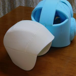 May include: Two pieces of protective headgear. One is a white, fitted cap with stitched seams. The other is a light blue, open-faced helmet made of foam, with a strap across the top and ear protection.