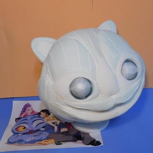 May include: A large, white cat head prop with mesh eyes and a detailed, stitched design. The prop is resting on a blue surface with a colorful illustration of a cartoon cat and people.