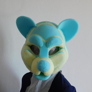 Könnte beinhalten: Eine blau-gelbe bärenartige Maske mit Netzkappen für die Augen. Die Maske hat große, abgerundete Ohren und eine markante Schnauze. Das Farbschema ist eine Kombination aus Hellblau und Gelb, wodurch ein verspieltes Aussehen entsteht. Die Maske wird von einer Person getragen.