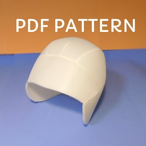 May include: A white, textured hat with a curved brim and ear flaps. The text "PDF PATTERN" is displayed in white block letters above the hat. The hat is set against a blue and orange background.