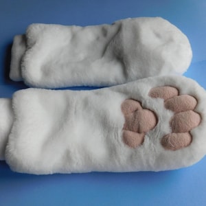 May include: A pair of white faux fur mittens designed to resemble animal paws. One mitten features a soft, light pink padded paw print detail on the palm side, while the other mitten is plain white faux fur on both sides.