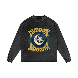 May include: A black long-sleeve shirt with a faded, vintage look. The front features a circular design with the word "HUZOOR" in a yellow, retro font above and below a crescent moon and star graphic. The design includes Arabic text.