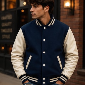 May include: A navy blue varsity jacket with cream-coloured sleeves, a ribbed collar, cuffs, and hem featuring white stripes. The jacket has a snap-button front closure, two side pockets with cream trim, and a logo at the bottom reading R LEATHERS.