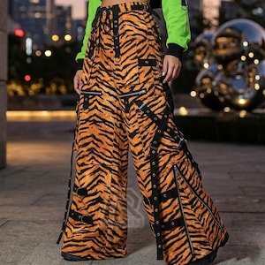 Könnte beinhalten: Orange-schwarze Cargo-Hose mit Tigerstreifenmuster mit mehreren Taschen, Reißverschlüssen und schwarzen Riemen mit Metallbeschlägen. Die Hose mit weitem Bein hat einen Kordelzug in der Taille.