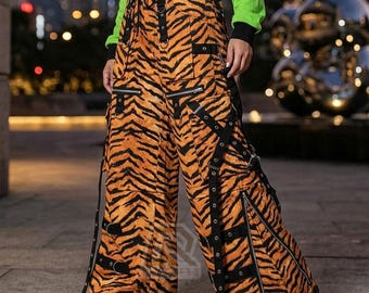 Op de afbeelding: Oranje en zwarte cargobroek met tijgerstreepmotief met meerdere zakken, ritsen en zwarte bandjes met metalen hardware. De broek met wijde pijpen heeft een taille met trekkoord.