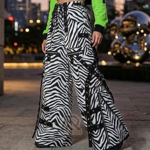 May include: Zebra print wide-leg trousers with black detailing, zips, and straps. The black and white pattern is striking. The trousers have a high waist and a drawstring closure.