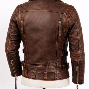 May include: A brown leather jacket with a distressed finish. The jacket features a notched collar, multiple zippered pockets, and ribbed detailing on the shoulders and elbows. It has a belt at the waist and a zipper closure. The jacket is displayed on a mannequin.