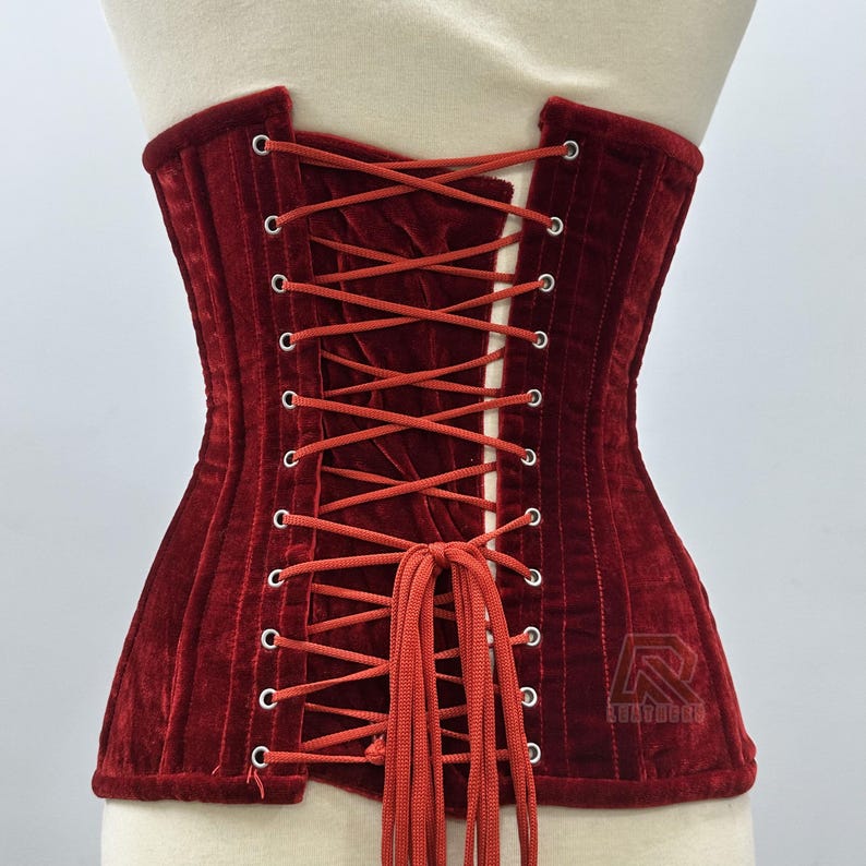 Puede incluir: Un cors&eacute; de terciopelo rojo oscuro con la espalda con cordones. El cors&eacute; tiene un dise&ntilde;o estructurado con costuras verticales y un panel blanco contrastante en la parte posterior. El cord&oacute;n es de color rojo a juego. El cors&eacute; se exhibe en un maniqu&iacute;.