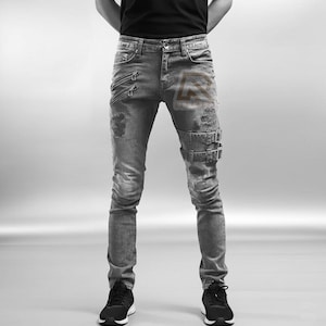 Może przedstawiać: Szare dżinsy z przetarciami, zamkami i sprzączkami. Jeansy mają dopasowany krój i są noszone z czarnymi trampkami. Ogólny styl jest odważny i nowoczesny.