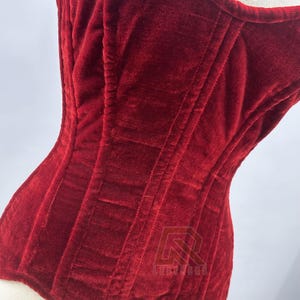 Puede incluir: Primer plano de un cors&eacute; de terciopelo rojo intenso. El cors&eacute; tiene costuras verticales y un dise&ntilde;o estructurado. La tela tiene un aspecto suave y texturizado. El cors&eacute; se muestra en un maniqu&iacute;.